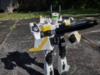 The VF-1S in battroid (robot) mode, posed and wielding its gunpod. The head of the mecha is white with a green visor and a red painted light on the top of the head and black antennae on the sides. Colors otherwise carry-over from fighter mode and is predominantly white, with the thighs being black.
